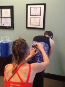woman with braided hair in red top with hands on the back of a client wearing a purple top that is bending over to check for scoliosis