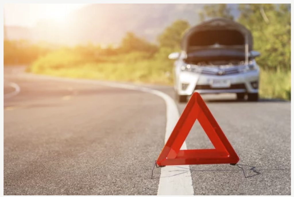 A car on the right side of the road is out of focus and has the hood open. In the foreground a change of focus happens on a red cone with an opening in the middle is upright on the line on the right side of the image. The sky is light on the upper left side of the image.