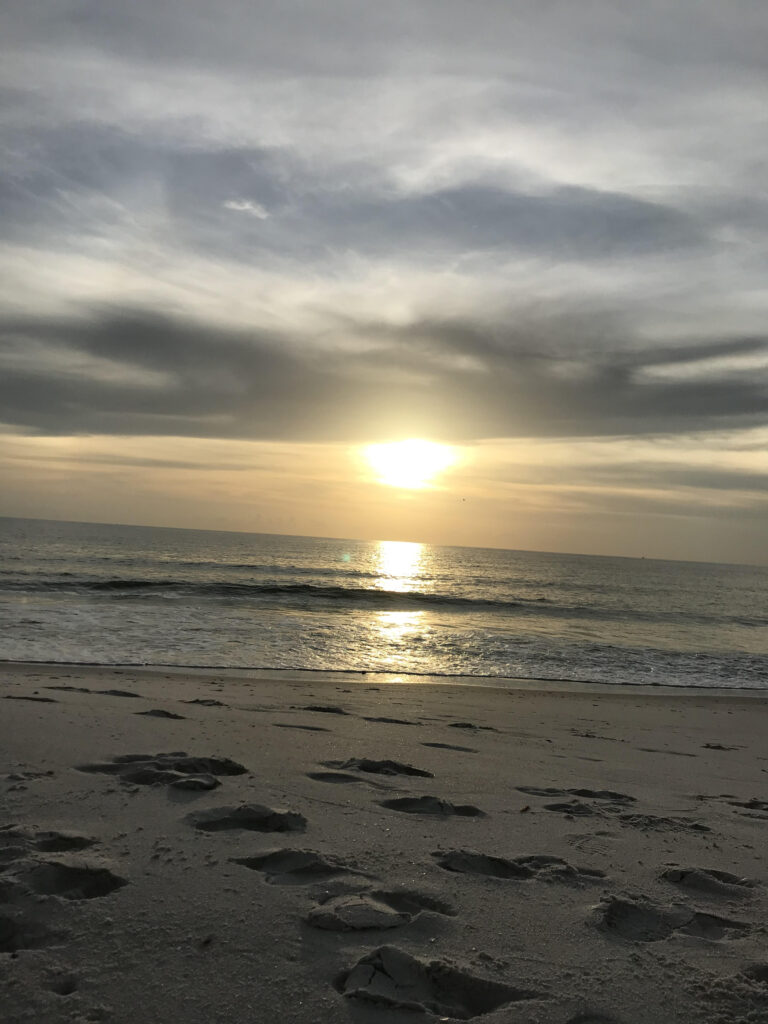 an overcast sky with an orange sun setting in the middle. water flows to the front of the picture and sand is in the foreground with small prints imprinted. A calm sense of hope and love is present.